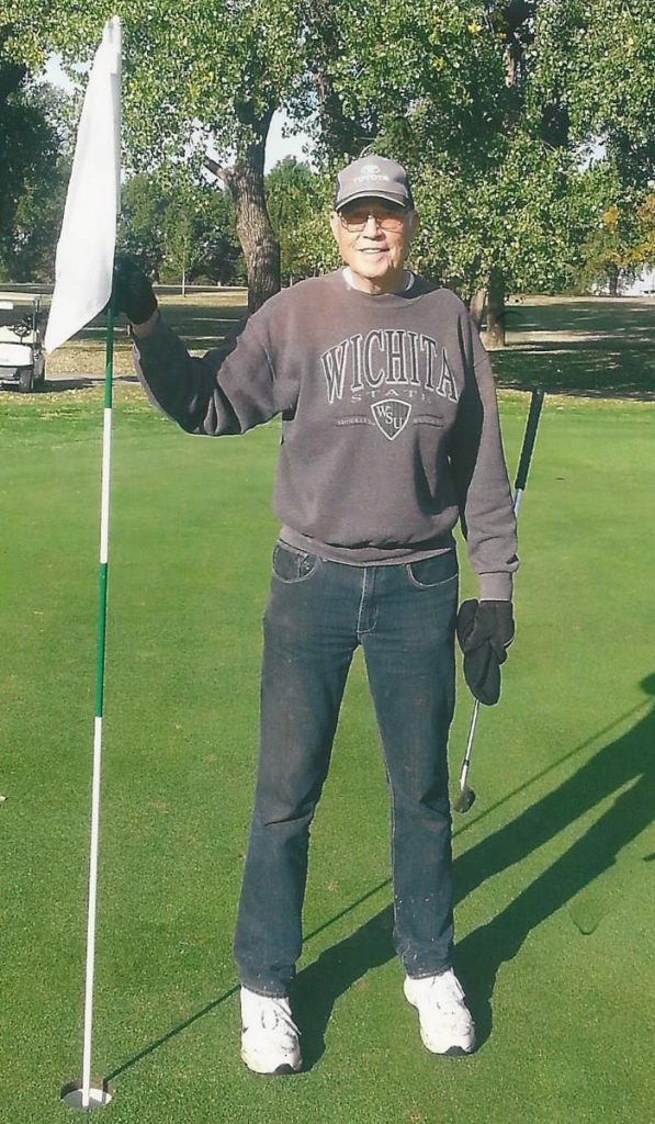 Man standing holding golf flag wearing Wichita State University sweatshirt and Toyota ball cap