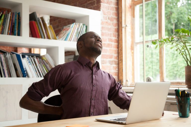 Young exhausted african american male office worker having back discomfort at work desk in office. Fatigued casual businessman feeling back pain after sitting on uncomfortable chair for long hours.