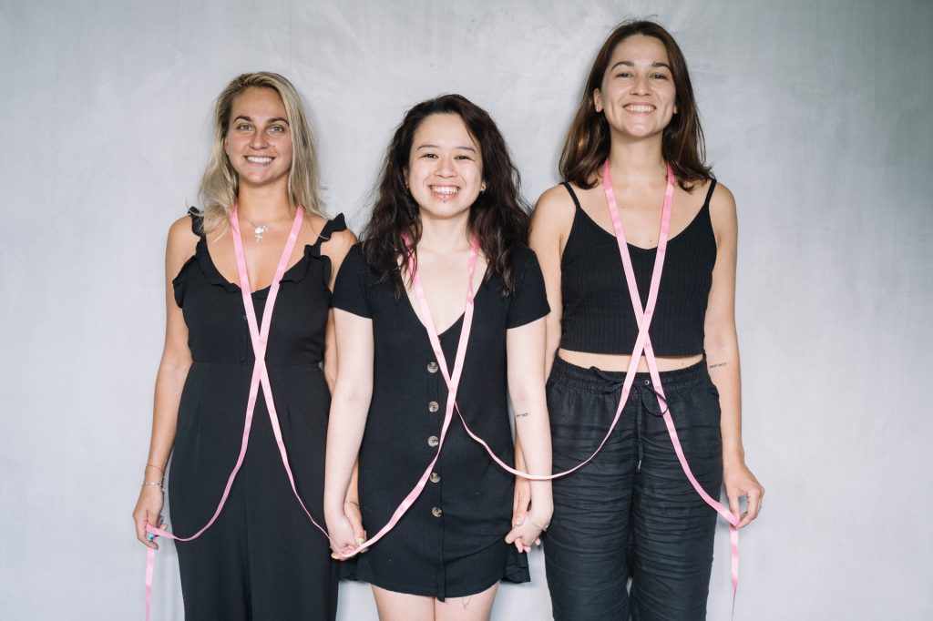 three women standing with pink ribbon around their neck holding hands in solidarity for breast cancer awareness