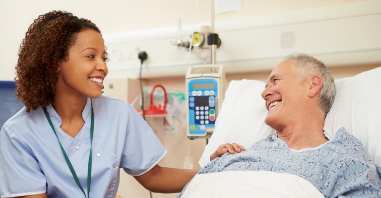 black woman nurse caring for white man patient in hospital bed - nurse has hand on patient's shoulder and smiling and talking about NMC Health hospital care with the patient portal