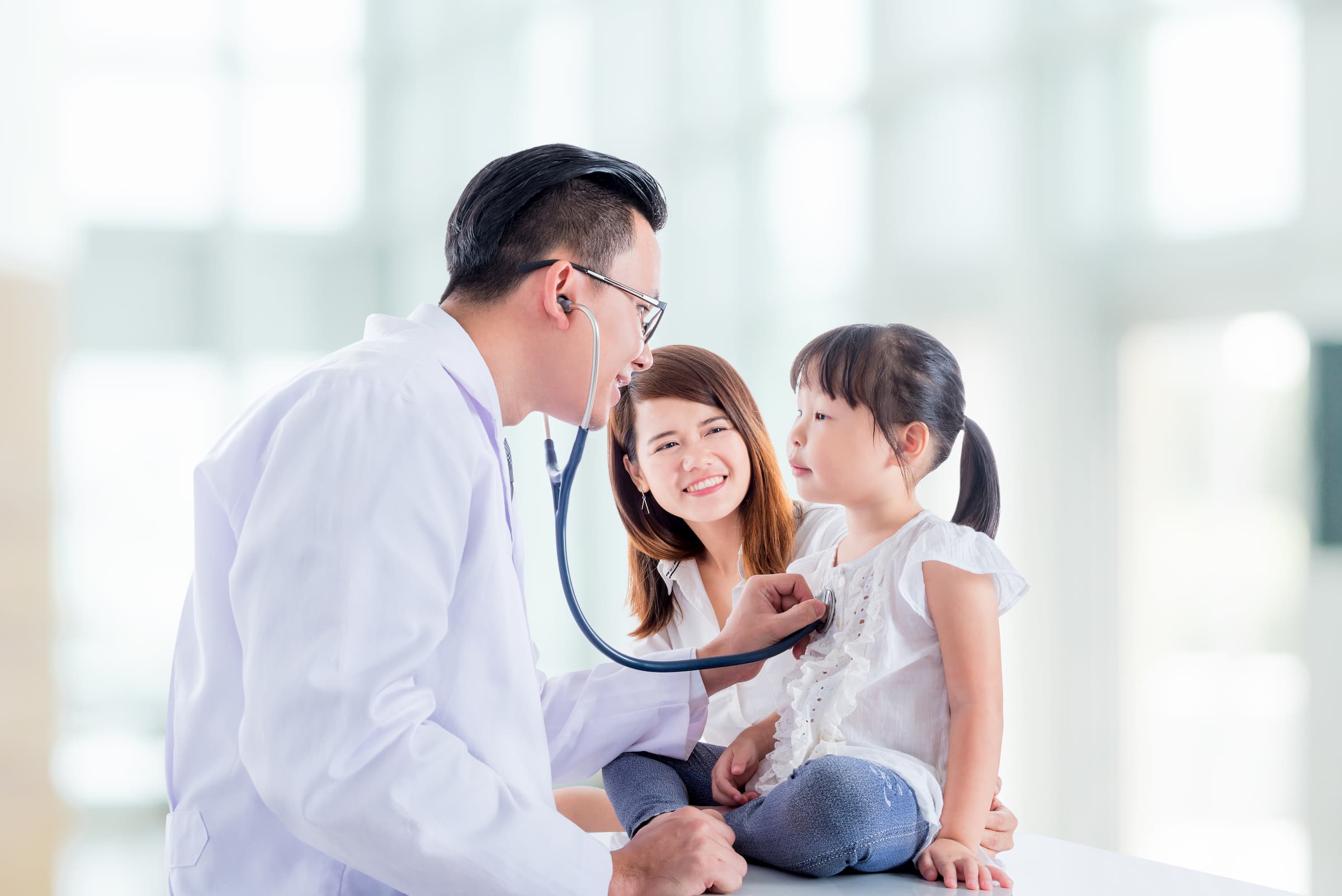 white doctor using blue stethoscope on asian girl as her mom looks at her and smiles