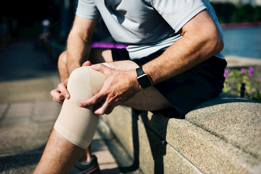 man sitting on curb rubbing left knee because knee pain, knee in knee brace joint pain