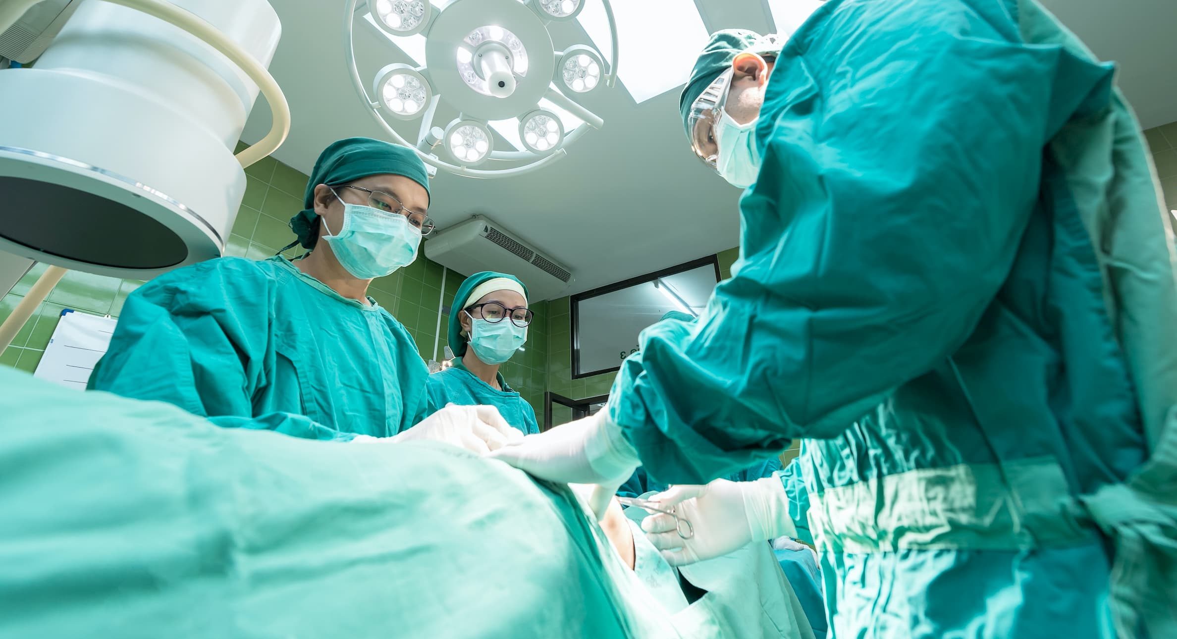 three doctors wearing aqua scrubs in surgery performing a procedure in an operating room on a patient who is laying on a table