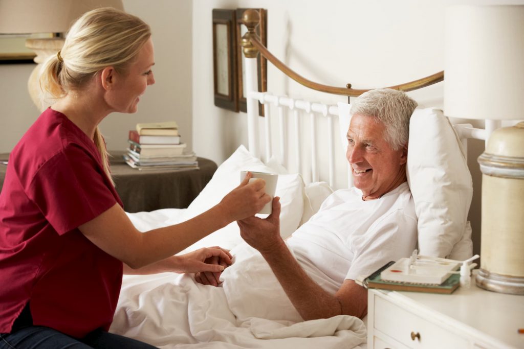 white caregiver helping white man laying in bed with getting a drink of coffee