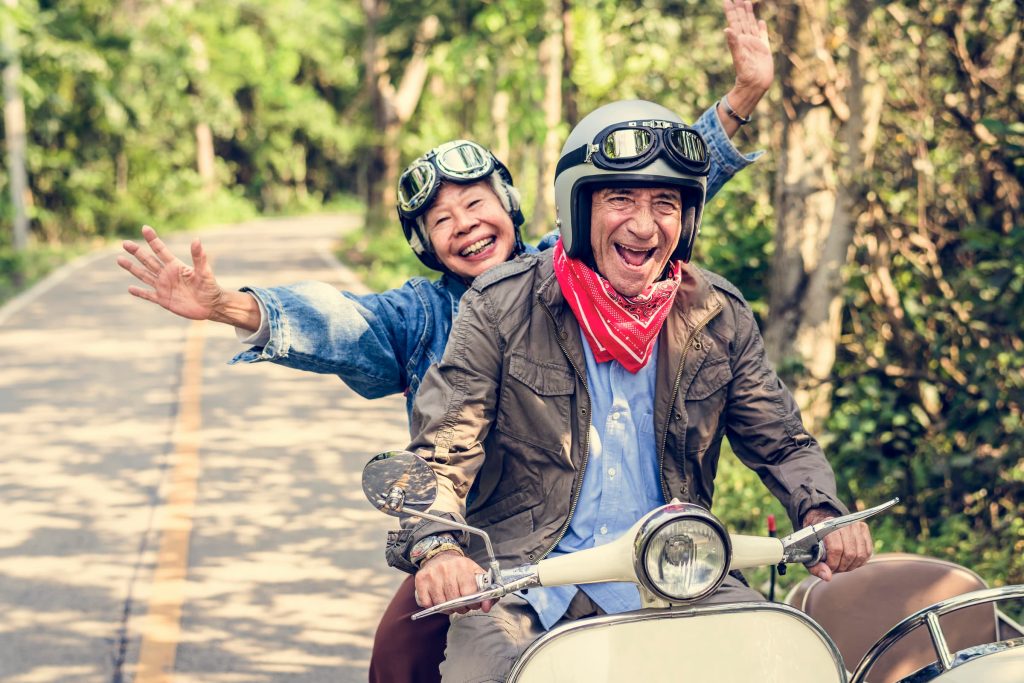 senior hispanic man and asian senior woman riding motorcycle with smiles on their face down wooded road with hands up