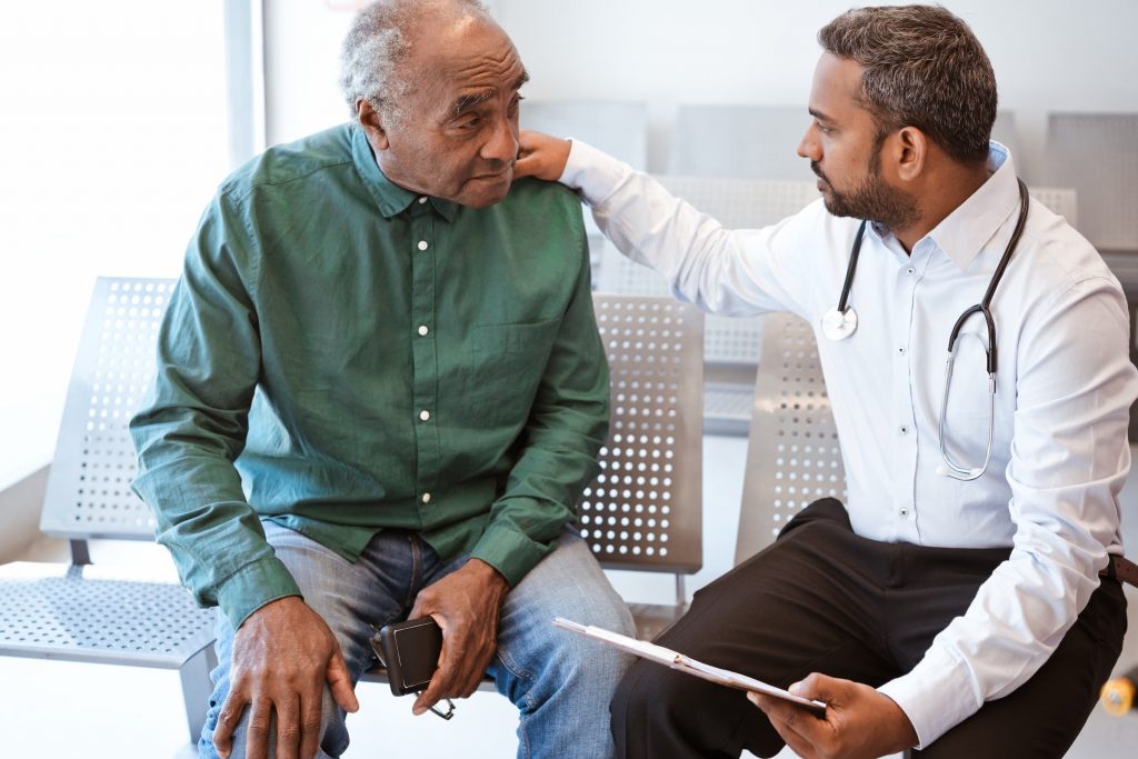 black senior seeing male doctor with hand on his shoulder sitting in patient room