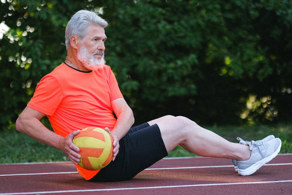 active white senior man in orange shirt and black shorts working out while sitting on outdoor track with exercise ball