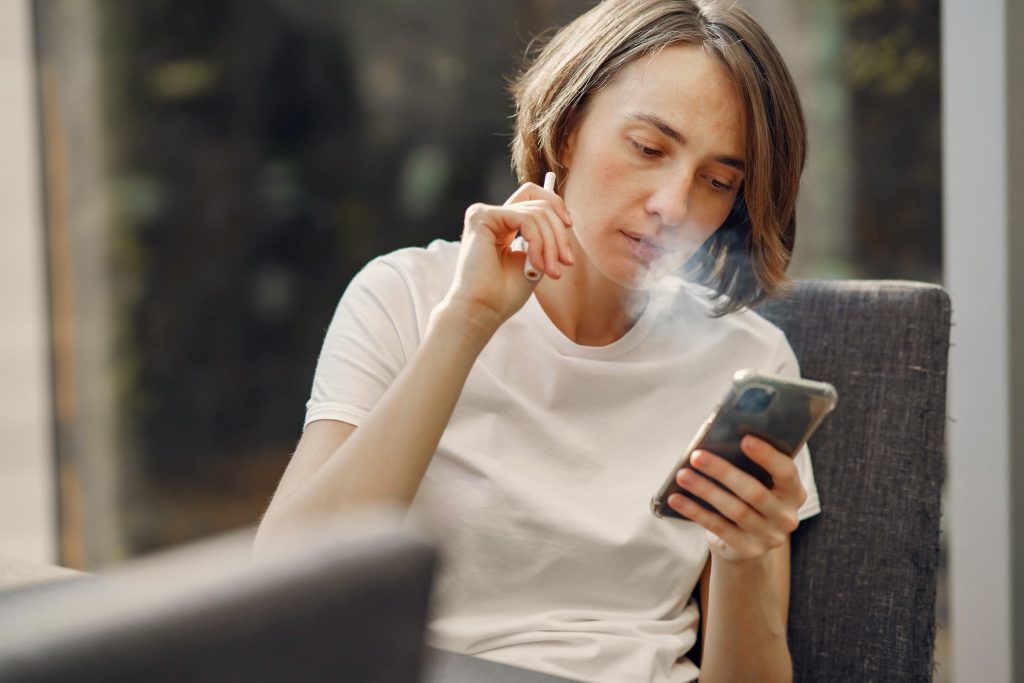 woman with short brown hair sits at table with phone in hand and vaping smoke e-cigarette blowing out smoke