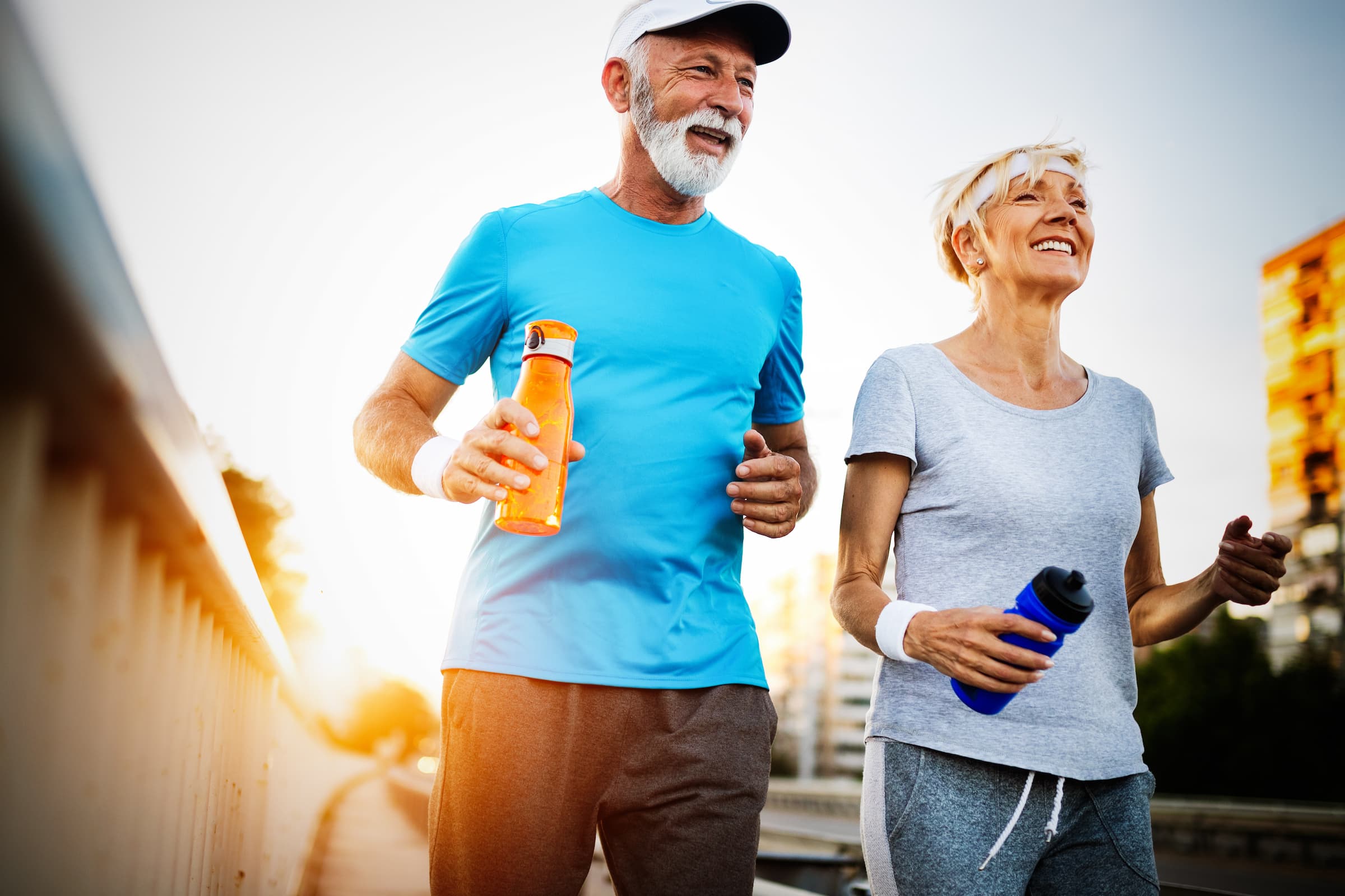 active seniors older man in blue shirt and yellow water bottle with wife who has blue water bottle and gray shirt smiling with sun setting behind them while they're walking outside
