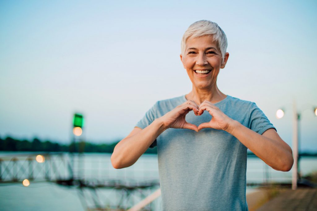 white elder woman with white hair making heart shaped hands on her chest wearing gray shirt standing outside next to water and street sign