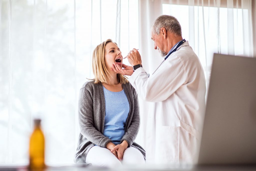 senior doctor examining a young woman in office for allergies looking at her throat as she sits on an exam table