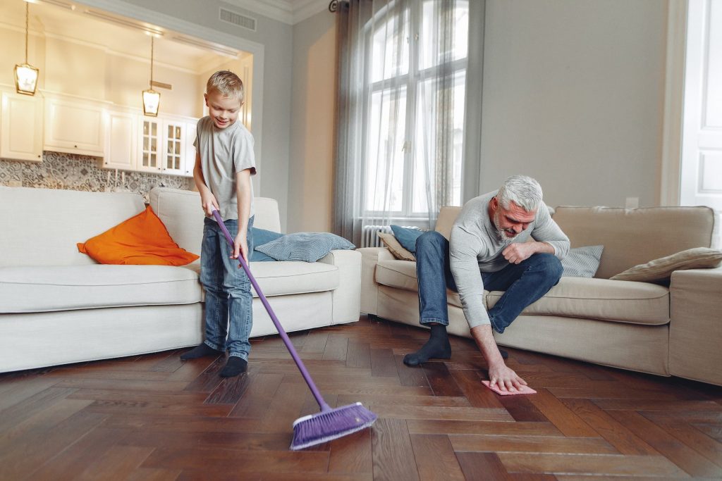 young boy and older man sweeping and cleaning wood floor in home sitting on white couches with orange and blue pillows, broom, mop, etc, clean for allergies