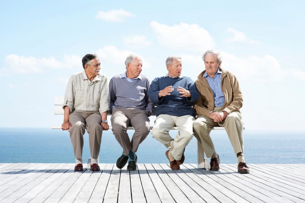 four senior men sitting on a bench on a dock with the ocean and sky behind them