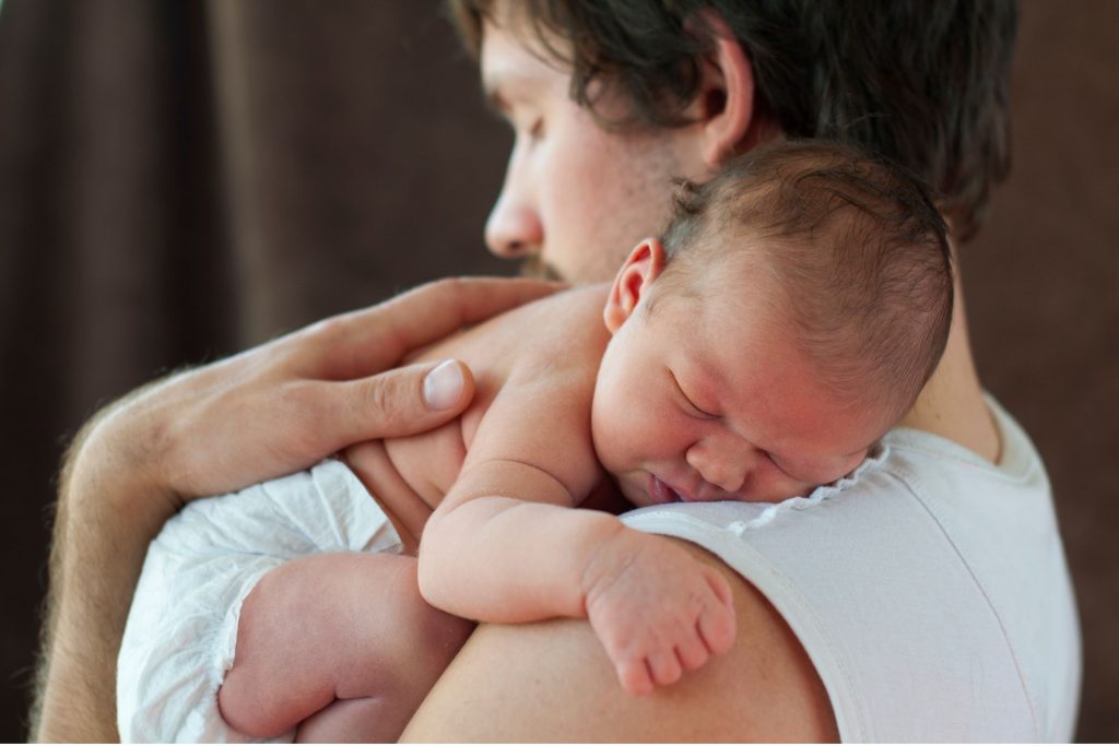 Young white dad holding newborn baby over left shoulder as baby sleeps