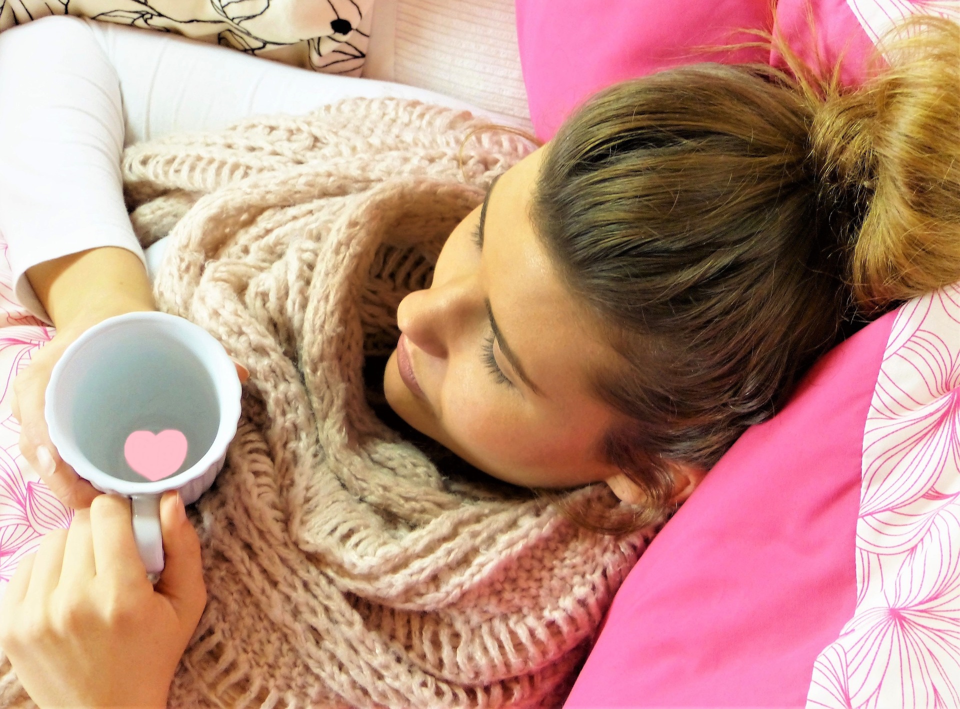 young woman in bed wrapped in warm blanket holding a cup of warm liquid, possibly ill or sick