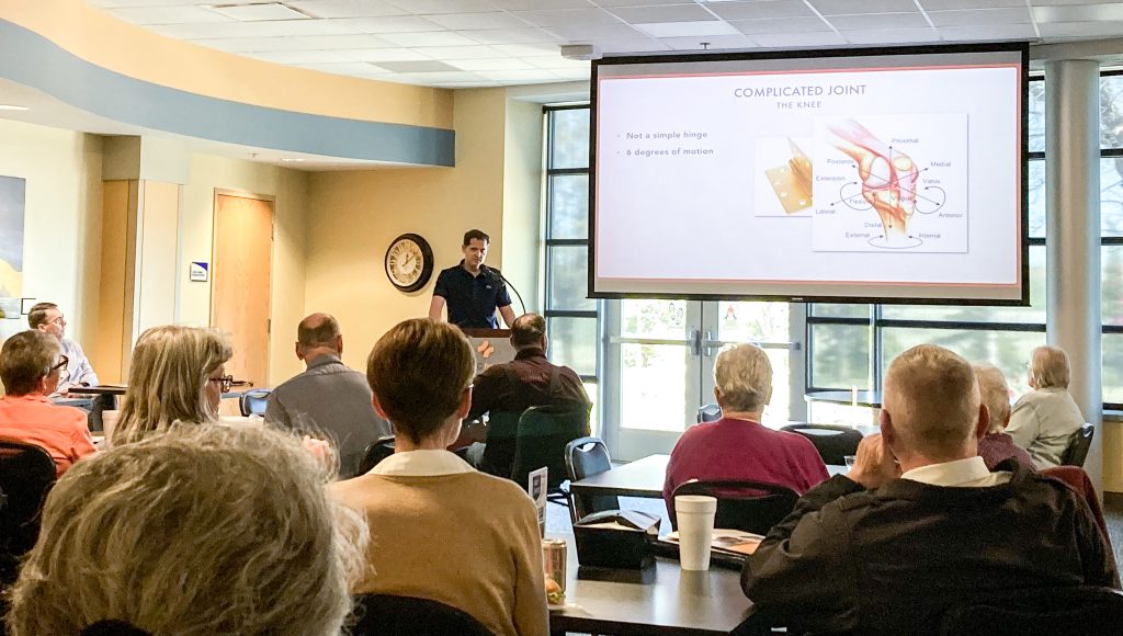 man at podium speaking at wellness first Wednesday to audience about wellness with large projection screen showing text joint pain as part of the classes and events at NMC health