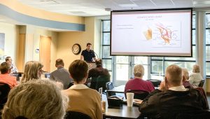 man at podium speaking at wellness first Wednesday to audience about wellness with large projection screen showing text joint pain as part of the classes and events at NMC health