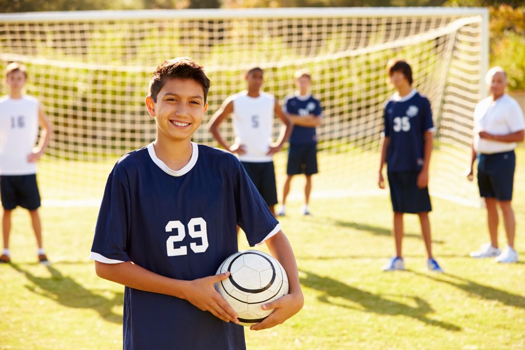 boys from two soccer teams facing the camera and smiling while on the soccer field in front of the goal, and one boy is holding the soccer ball