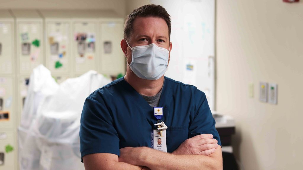man wearing a COVID mask in scrubs looking at the camera with folded arms