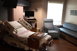 An alternative view of the birthing suite, highlighting a long cushioned bench seat (sofa bed) under a large window, intended for a partner or family member to stay overnight. The room is softly lit, emphasizing a private and calm environment.