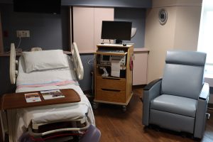 A hospital delivery room featuring a modern medical bed with a white pillow and pink-and-blue striped blanket. To the right is a wooden medical cart with a monitor and fetal tracking equipment, alongside a blue reclining chair for visitors.