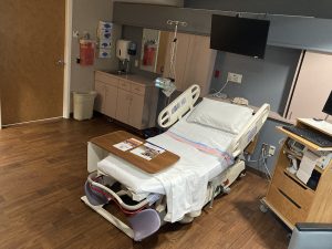 A wide-angle view of the birthing suite showing the adjustable medical bed, a sink area with medical supply dispensers, and a wall-mounted television. The room has warm wood-laminate flooring and neutral-toned cabinetry.