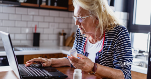 elderly woman holding prescription medicine bottle in one hand while looking at her laptop