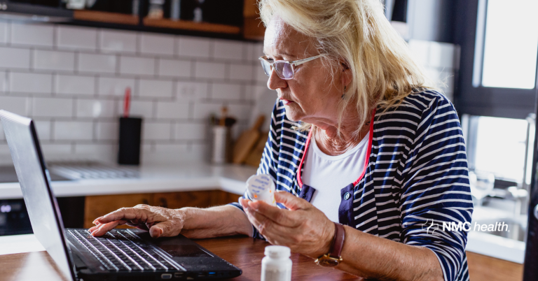 elderly woman holding prescription medicine bottle in one hand while looking at her laptop