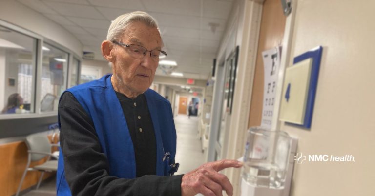 older gentleman in hospital hallway pointing to hand sanitizer station wearing blue volunteer vest
