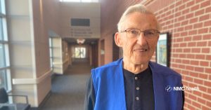 older smiling man standing in hospital hallway wearing blue volunteer vest