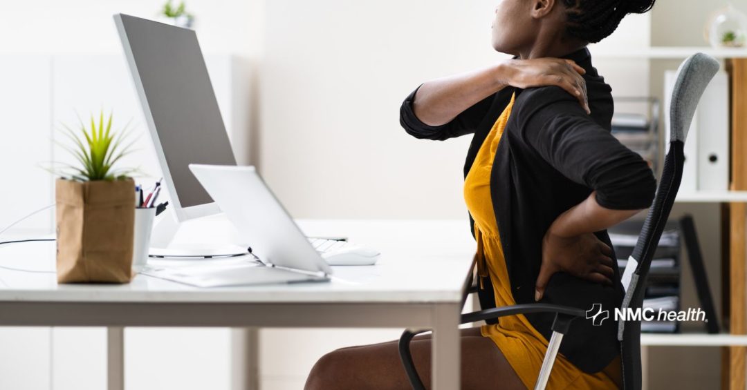 professional african american woman at desk with one hand on her lower back and the other hand on her shoulder