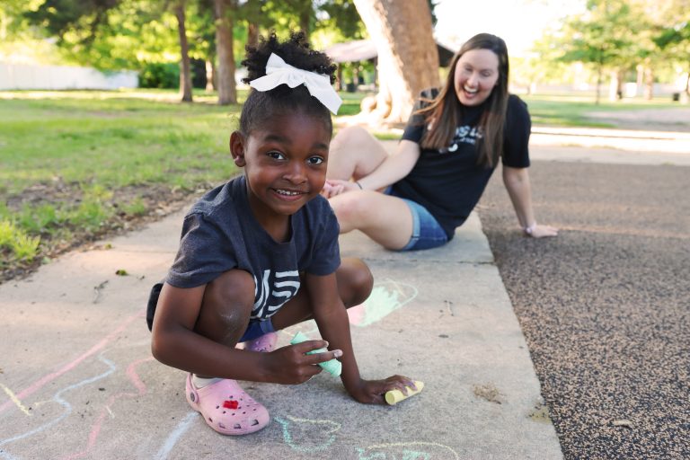 young african american girl smiling and playing with sidewalk chalk with smiling female in the background