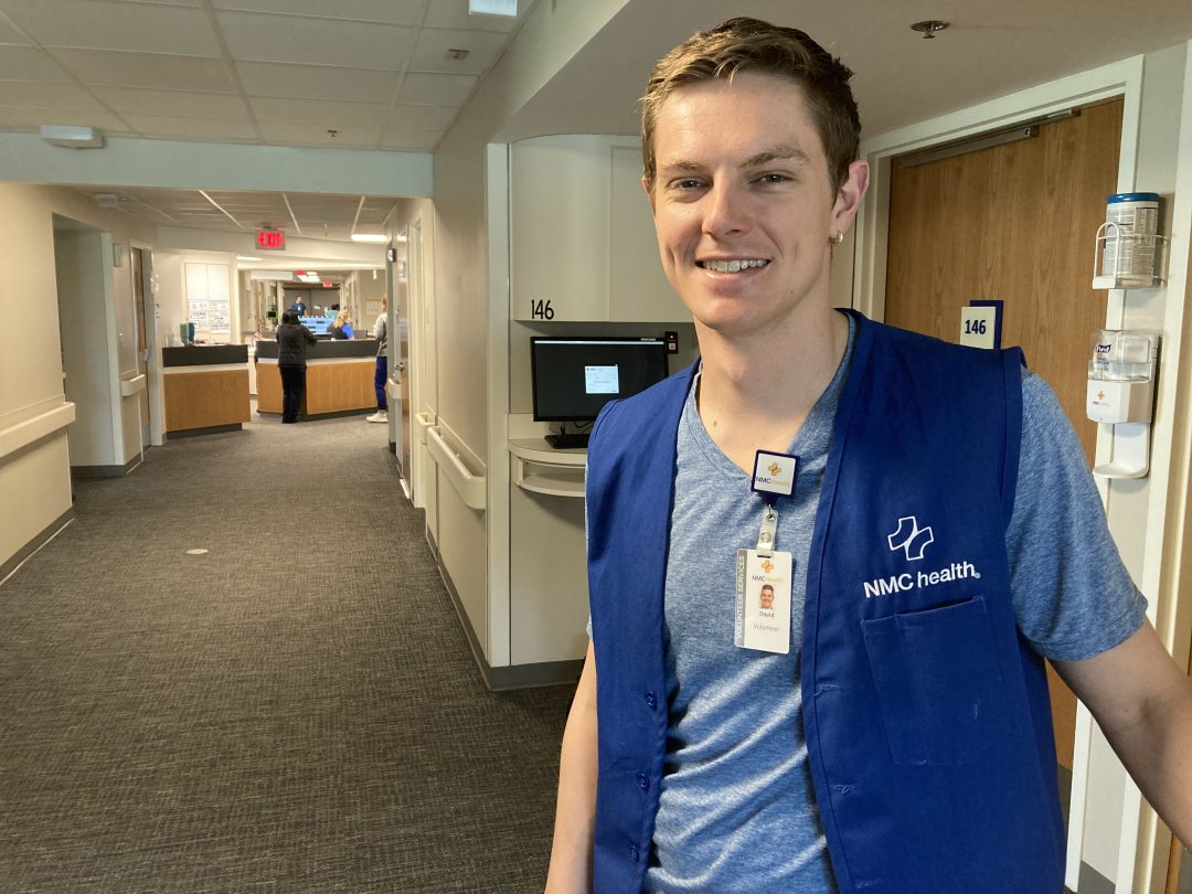 young man smiling and standing in hospital hallway wearing blue vest