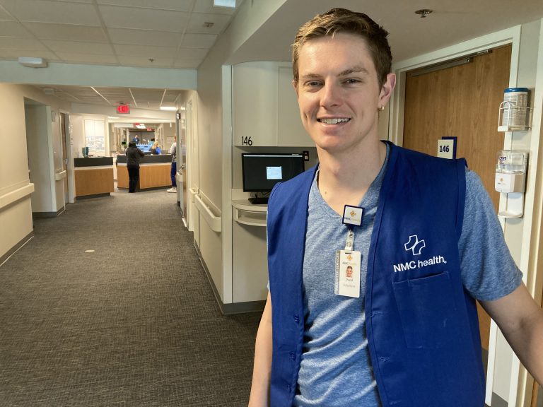 young man smiling and standing in hospital hallway wearing blue vest