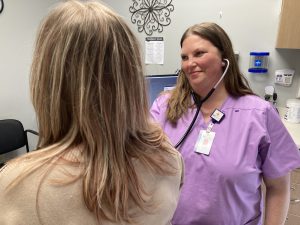 smiling healthcare provider wearing purple scrubs standing next to patient while listening to stethoscope