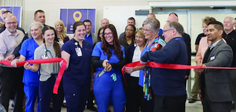 medical staff and community members surrounding nurse in blue scrubs holding large pair of scissors after ribbon is cut
