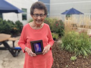 elderly woman outside smiling at camera holding picture of elderly man
