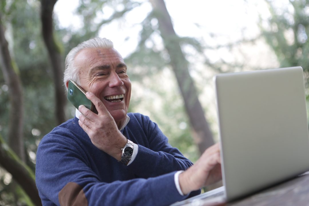 senior white man smiling and talking on phone and playing on laptop medicare open enrollment
