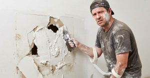 man covered in spackle attempting to patch large hole in wall