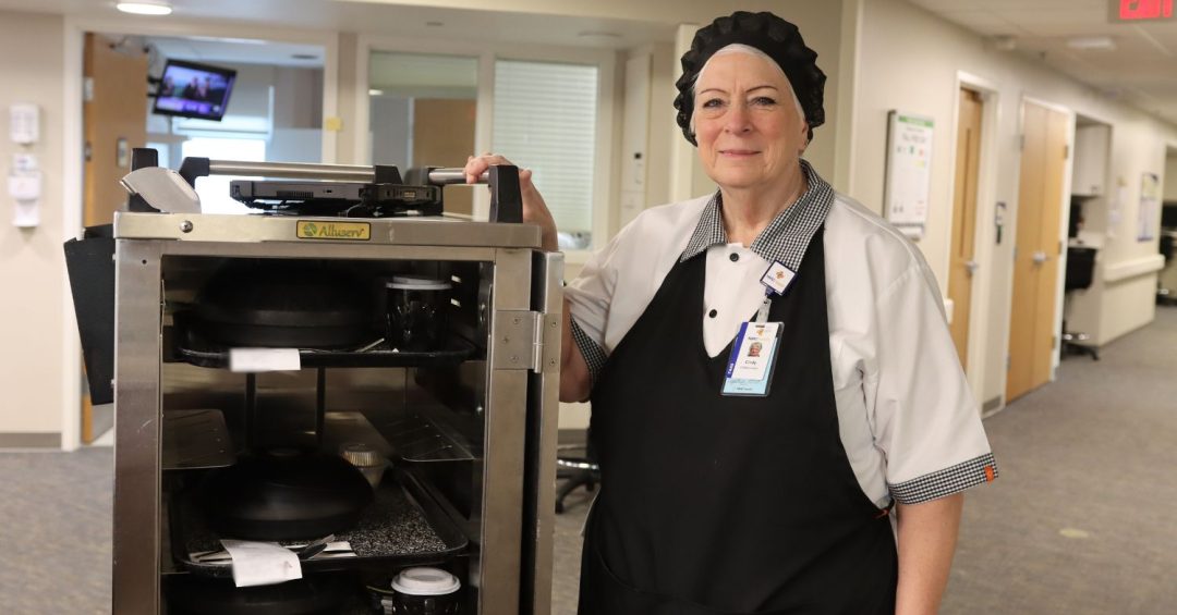 a woman in a black apron and black hat standing next to a metal cart