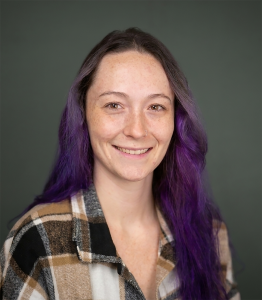 Woman with purple-dyed hair smiling for a headshot in front of a grey backdrop