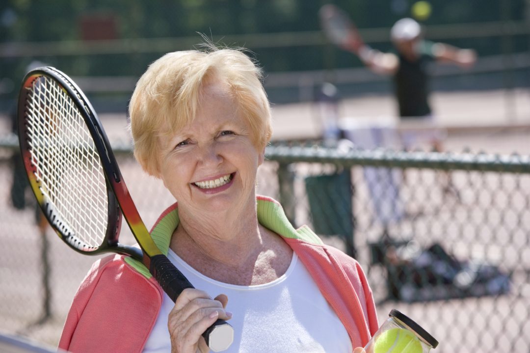 smiling older woman on tennis court holding tennis racket and tennis ball