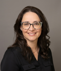 Woman with dark brown hair and cheetah print glasses wearing a black blouse and posing for a headshot in font of a grey backdrop