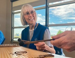 A woman wearing glasses and a blue vest over a striped shirt smiles while holding mallets to play a harp.