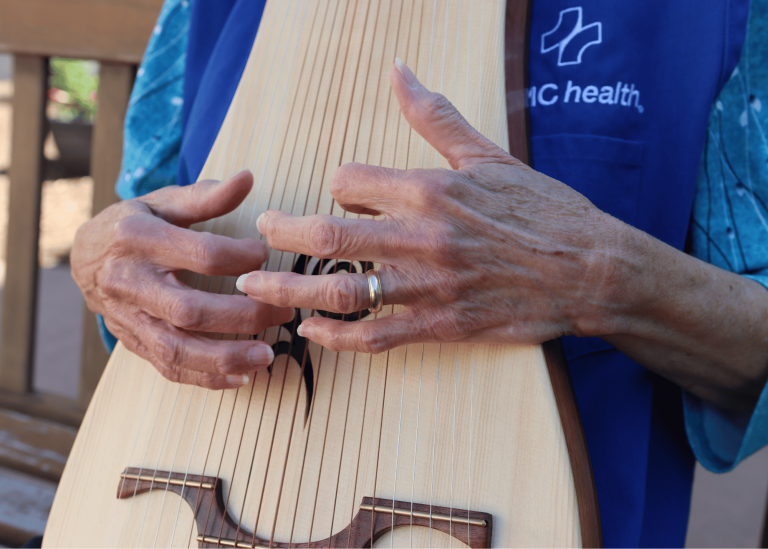 Close-up of hands playing a stringed instrument with many strings and a wooden body. The person wears a blue vest with a white health logo and text.