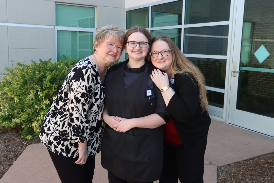 Three people smiling at the camera. The person in the middle wears a black apron, and the person on the right wears a black blazer and a gold watch.